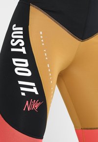 Leggings de sport noirs, dorés et rouges avec une texture lisse ; arborent "JUST DO IT" en blanc et le logo en rose près de l'ourlet.