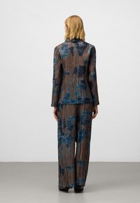 Vrouw met blond haar draagt een bruin en blauw bloemenplooisuit met wijde broeken, staande met de rug naar de camera op een effen grijze vloer.