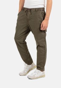 Man wearing olive green drawstring pants and white athletic sneakers, standing with hands relaxed at sides.