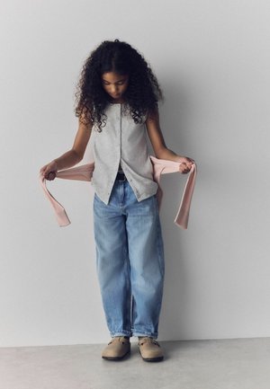 Mladá dívka s kudrnatými vlasy se dívá dolů, zatímco drží růžový svetr, má na sobě šedý rozepnutý top, volné džíny a béžové boty.