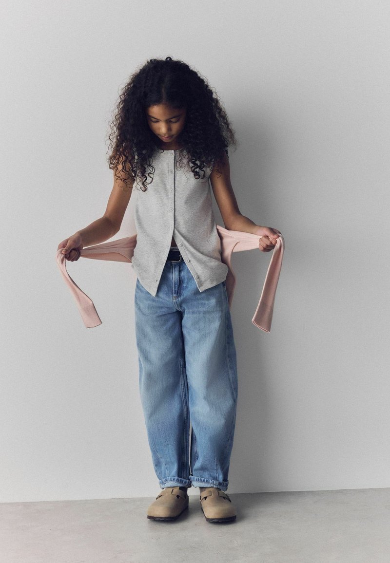 Jeune fille aux cheveux bouclés regardant vers le bas tout en tenant un pull rose, portant un haut gris à boutons, un jean ample et des chaussures beiges.