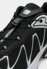 Sapatilha atlética preta com cabedal em malha e sintético, detalhes prateados, atacadores elásticos e fecho por toggle. Solado robusto com padrão texturizado.