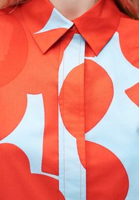 Κόκκινη μπλούζα με μεγάλα ανοιχτόγαλάζια αφηρημένα λουλουδένια σχέδια και μυτερό γιακά, κουμπωμένη στο λαιμό.