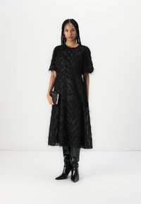 Robe midi noire à plumes avec des manches courtes, ondulations texturées et une silhouette évasée. Associée à des bottes noires jusqu'aux genoux et une pochette.