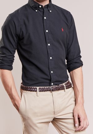 Homme portant une chemise noire boutonnée avec les manches retroussées, un pantalon beige et une ceinture en cuir marron tressé, debout avec une main dans la poche.