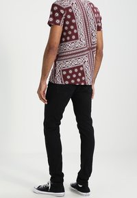 Krótki rękaw bordowej koszuli z białymi wzorami paisley i geometrycznymi, w połączeniu z czarnymi dżinsami skinny i czarnymi sneakerami, widok z tyłu.