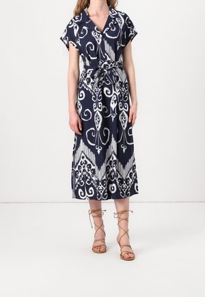 Femme portant une robe midi à motifs bleu marine et blanc avec manches courtes et taille nouée, associée à des sandales à brides couleur sable.