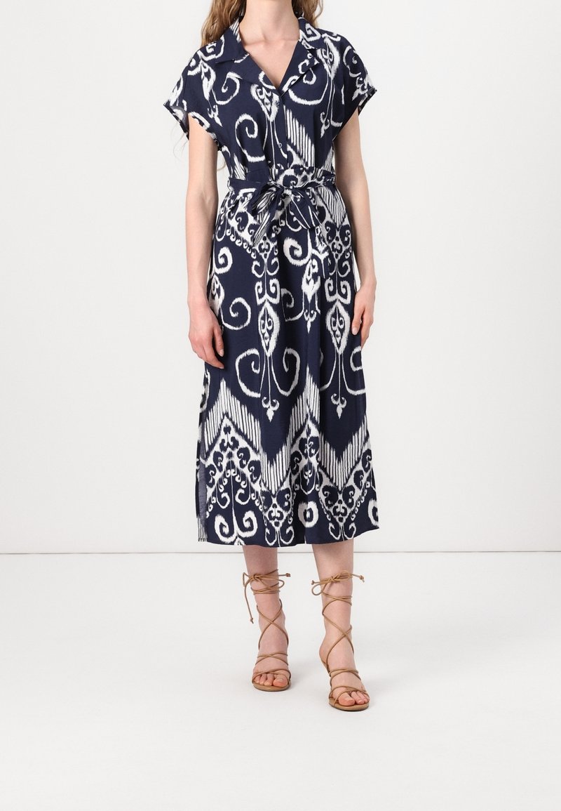 Femme portant une robe midi à motifs bleu marine et blanc avec manches courtes et taille nouée, associée à des sandales à brides couleur sable.