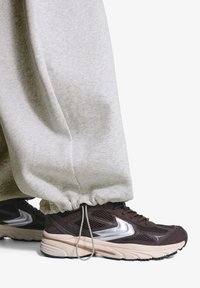 Brun træningssko med sølvaccenter og en tekstureret overdel, parret med lys grå, opfoldede sweatpants lavet af blødt materiale.