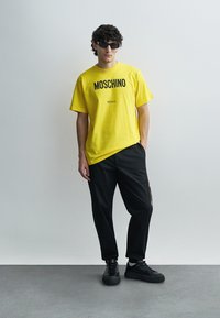 Gele katoenen T-shirt met zwarte "Moschino" branding, gecombineerd met zwarte taps toelopende broek en zwarte sneakers. Gladde texturen, casual ontwerp.