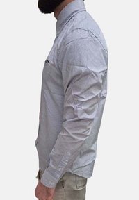 Camicia bianca con motivo a quadri blu, maniche lunghe, colletto a punta e taschino sul petto. Il tessuto appare liscio con una leggera lucentezza.