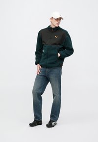 Tummanvihreä fleecevetoketjullinen takki, jossa musta kangaspaneeli ja oranssi logo; yhdistetty sinisiin farkkuihin ja mustiin kenkiin, joissa on harmaita yksityiskohtia.