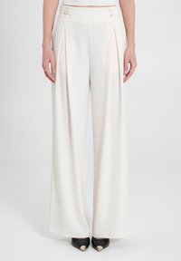 Pantaloni a gamba larga in tessuto crema con pieghe frontali, caratterizzati da elementi in oro alla vita. Tacchi neri in camoscio con accenti dorati completano l'outfit.