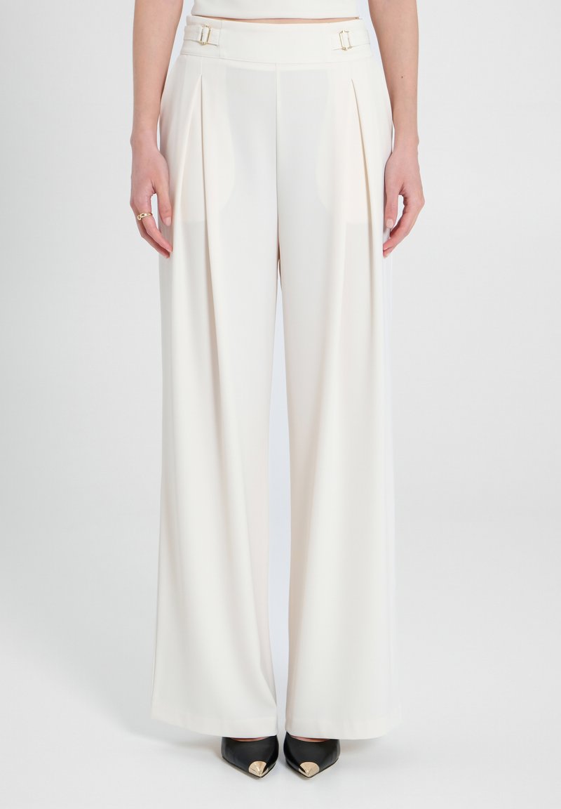 Pantaloni a gamba larga in tessuto crema con pieghe frontali, caratterizzati da elementi in oro alla vita. Tacchi neri in camoscio con accenti dorati completano l'outfit.