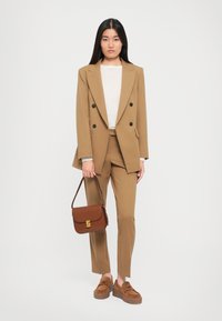 Brun skreddersydd dress med dobbeltspent jakke, lys beige genser, brun håndveske med gull detaljer, og platåsko.