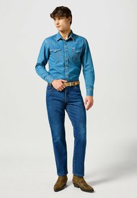 Camicia di jeans azzurro chiaro con tasche e cuciture a contrasto, abbinata a jeans blu scuro e stivali in pelle marrone. Vestibilità casual.