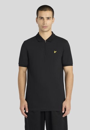 Homme debout face à l’avant, portant un polo noir à manches courtes avec un logo d’oiseau jaune sur la poitrine et une fermeture éclair partielle sur le devant.