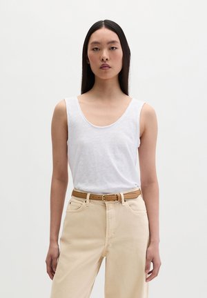 Giovane donna con capelli neri lisci che indossa una canottiera bianca, pantaloni beige a vita alta e una cintura marrone, in piedi su sfondo bianco.