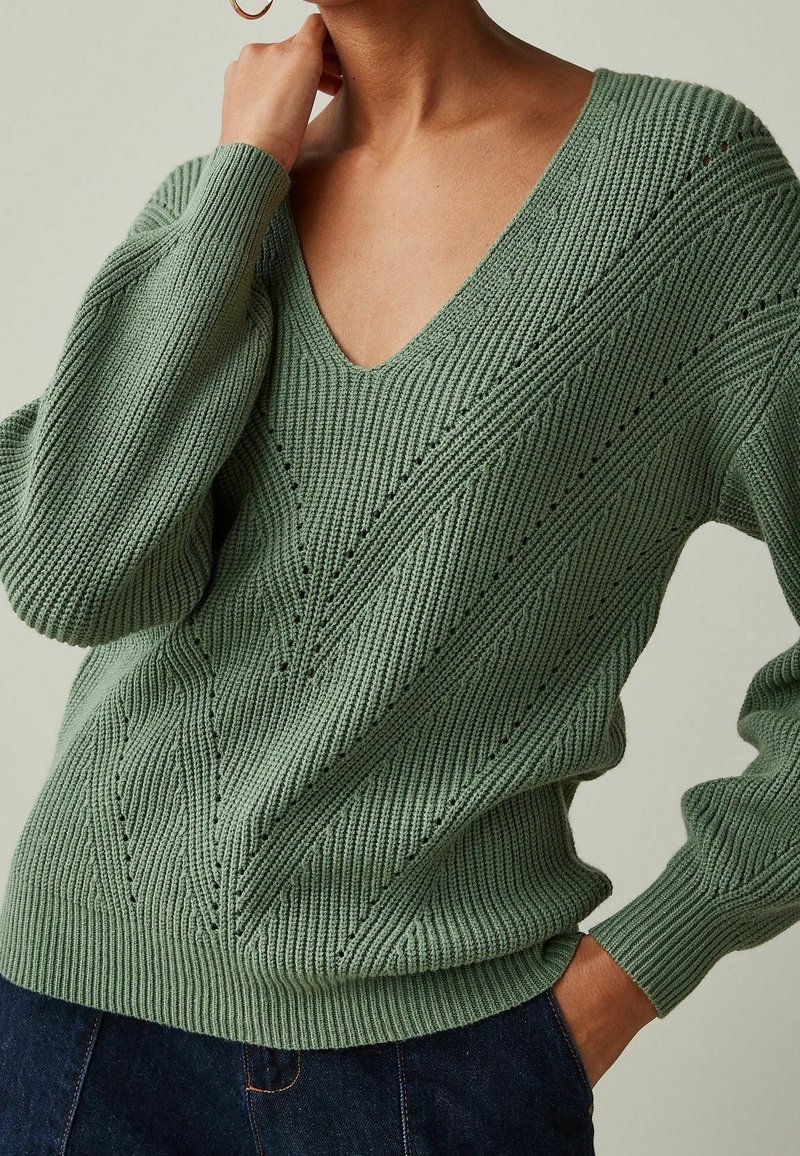 Pull en maille vert clair avec col en V, motif texturé et poignets en côtes. Présente une coupe décontractée et des détails de coutures visibles sur le corps.