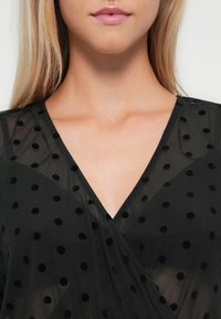 Zwarte doorzichtige stoffen top met een V-hals, versierd met verhoogde zwarte stippen, met een gladde textuur en gedrapeerd ontwerpelement.