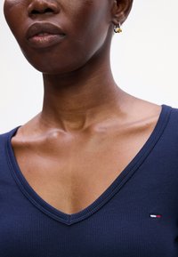 Top navy cu guler în V, cu mâneci scurte, având un mic logo în roșu, alb și albastru pe pieptul stâng și un material neted.