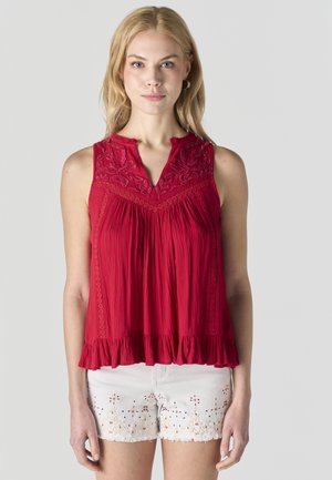 Mujer rubia vistiendo una blusa sin mangas roja bordada con bajo con volantes y pantalones cortos blancos con diseños florales de encaje calado.