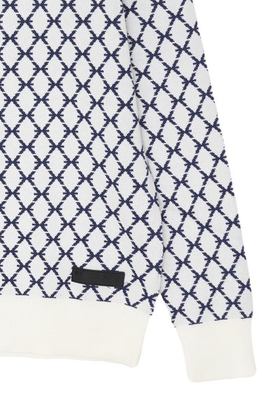 Manica di un maglione bianco con motivo a losanghe blu navy e polsino a costine, con un piccolo etichetta quadrata nera del marchio vicino all'orlo.