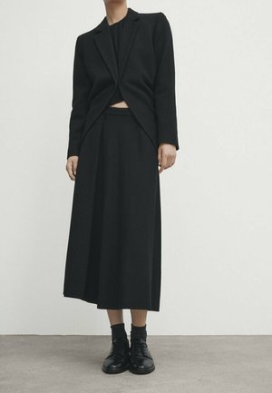 Personne portant une veste courte noire par-dessus un haut noir, une jupe midi noire à jambes larges, des chaussettes noires et des chaussures noires à lacets, debout sur un sol gris.