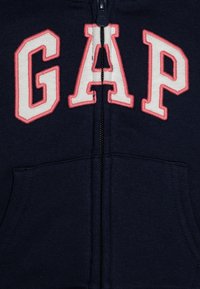 Hanorac închis la culoare, cu fermoar, cu un logo mare brodat în roz "GAP". Dispune de un fermoar central și două buzunare frontale. Material moale, texturat.