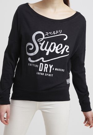 Woman wearing black off-shoulder sweatshirt with white "Super Dry Cutters & Makers Japan Spirit" text and white pants against gray background.