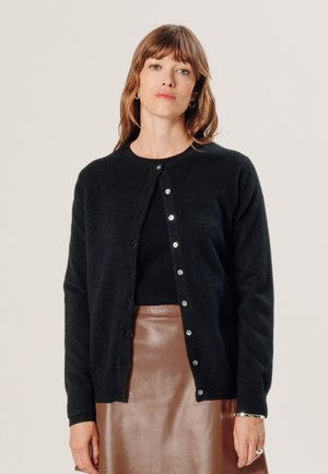 Schwarze Strickjacke aus weichem Material, mit rundem Halsausschnitt, durchgehender Knopfleiste und langen Ärmeln, kombiniert mit einem braunen Lederrock.