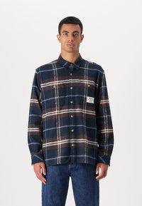 Camisa xadrez azul-marinho com riscas castanhas e brancas, design de botões, dois bolsos no peito e um rótulo tecido do lado esquerdo.