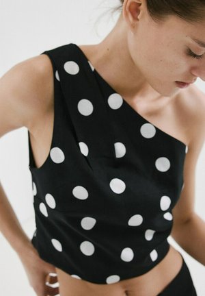 Femme portant un haut noir asymétrique à gros pois blancs, regardant vers le bas avec les mains posées sur les hanches.