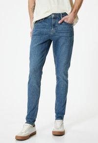 Jeans in denim azzurro chiaro con una vestibilità slim, dotati di cinque tasche, chiusura con zip e dettagli di cucitura sottili. Indossati con scarpe da ginnastica bianche.
