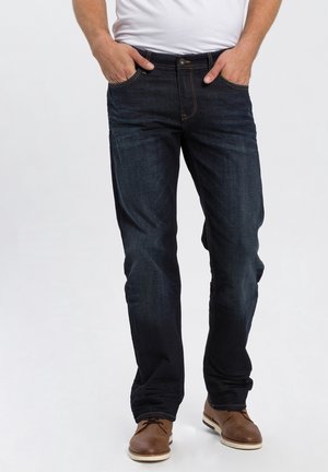 Homme portant un jean bleu foncé, les mains dans les poches, et des chaussures à lacets marron clair, debout sur un fond blanc uni.