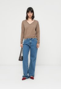 Maglione marrone a scollo a V, jeans azzurri a gamba larga, scarpe rosse, tiene una piccola borsa nera. Sfondo semplice senza elementi aggiuntivi.