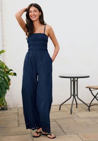 Mono azul marino con corpiño fruncido, piernas anchas y ribete de encaje en el dobladillo. Cuenta con tirantes ajustables y bolsillos. Se lleva con chanclas.