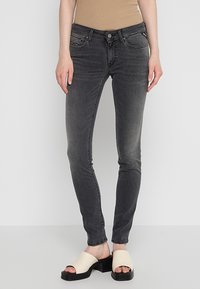 Jeans negros slim fit con un acabado desgastado, que presentan un diseño clásico de cinco bolsillos y detalles de herrajes metálicos. Combinados con sandalias deslizantes de color claro.