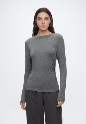 Femme aux longs cheveux foncés portant un haut gris ajusté à manches longues et un pantalon ample foncé, debout devant un fond blanc uni.