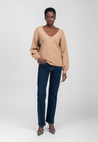 Beige gebreide trui met een diepe V-hals, losse pasvorm en lange mouwen, gecombineerd met donkerblauwe rechte jeans en zwart-wit gestreepte hakken.