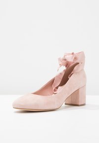 Růžová semišová bota na blokovém podpatku s kulatou špičkou a stuhou detail na kotníku. Měkká textura a hladký povrch s pevným základem.