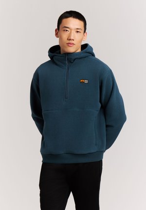 Jonge man met kort zwart haar die een blauwe fleece trui met capuchon en rits aan de voorkant en een zwarte broek draagt, staand tegen een effen achtergrond.