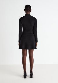 Abito nero a maniche lunghe con un corpetto aderente e gonna svasata. Tessuto a maglia liscio, colletto alto e lunghezza fino a metà coscia. Scarpe nere con tacco.