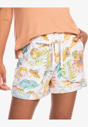 Personne portant un short blanc à cordon avec un imprimé floral tropical coloré et un haut pêche à manches courtes, main dans la poche, tatouage visible sur le bras.