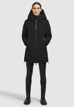 Black quilted puffer coat with a high collar and oversized hood. Features a front zipper and side pockets. Worn with black leggings and boots.