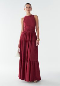 Kvinde iført en ærmeløs, tekstureret burgunder maxi kjole med samlet talje og lagdelt nederdel, der holder en lille hvid håndtaske, står på hvid baggrund.