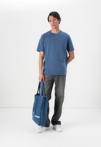 Camiseta de algodón azul con mangas cortas, pantalones de mezclilla grises y una bolsa tote de lona azul con texto blanco. El modelo está de pie contra un fondo liso.