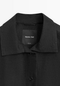 Schwarzes Hemd mit Kragen und Knopfverschluss, strukturierter Stoff und sichtbarem Innenetikett mit der Aufschrift "Massimo Dutti, Hergestellt in China."