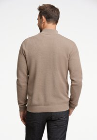 Suéter marrón de punto con una superficie texturizada, puños y dobladillo acanalados, con un cuello alto. Llevado con jeans gris oscuro.