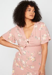 Różowa sukienka w kwiaty z krótkim rękawem, głębokim dekoltem w kształcie V i detalem w postaci zawiązania z przodu. Wykonana z lekkiego, gładkiego materiału w małe białe i żółte kwiatki.